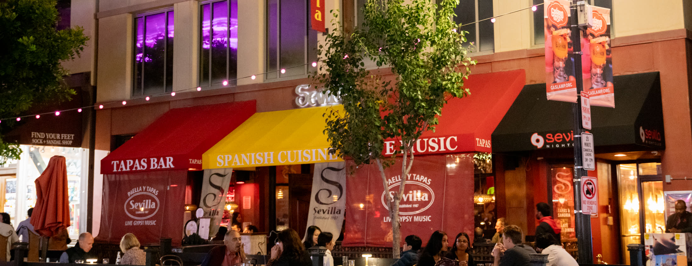 Caf&eacute; Sevilla&rsquo;s Gaslamp block at night: striped awnings, string lights and a full patio of guests
